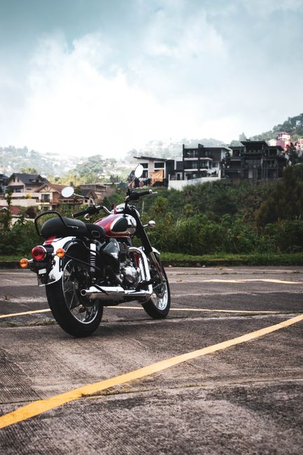 A black and chrome motorcycle parked diagonally in an outdoor parking lot with yellow parking lines, positioned with its rear facing slightly to the left and its front pointing towards the right. The motorcycle features a comfortable black seat, round rearview mirrors, and orange indicators. It is situated near the edge of the parking area, with a background of greenery, shrubbery, and modern residential buildings on a hillside. The sky above is partly cloudy with patches of blue, and the overall scene is well-lit with natural daylight. This image illustrates the type of vehicle that might be transported during home relocation or moving services offered by Man and Van Enfield, highlighting the transportation of personal belongings or vehicles as part of the moving process.