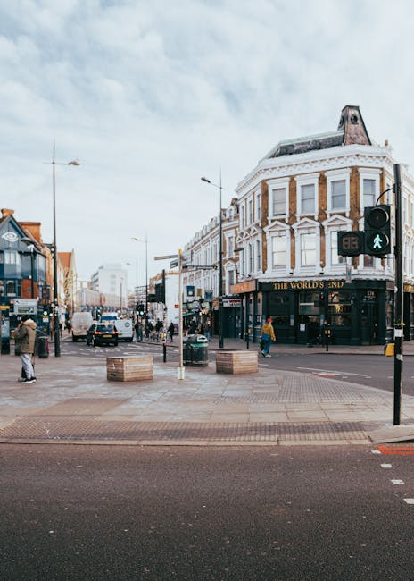A busy street scene in Enfield with a corner building featuring a white façade and multiple large windows, housing a pub called 'The World's End.' The street is filled with pedestrians waiting at the