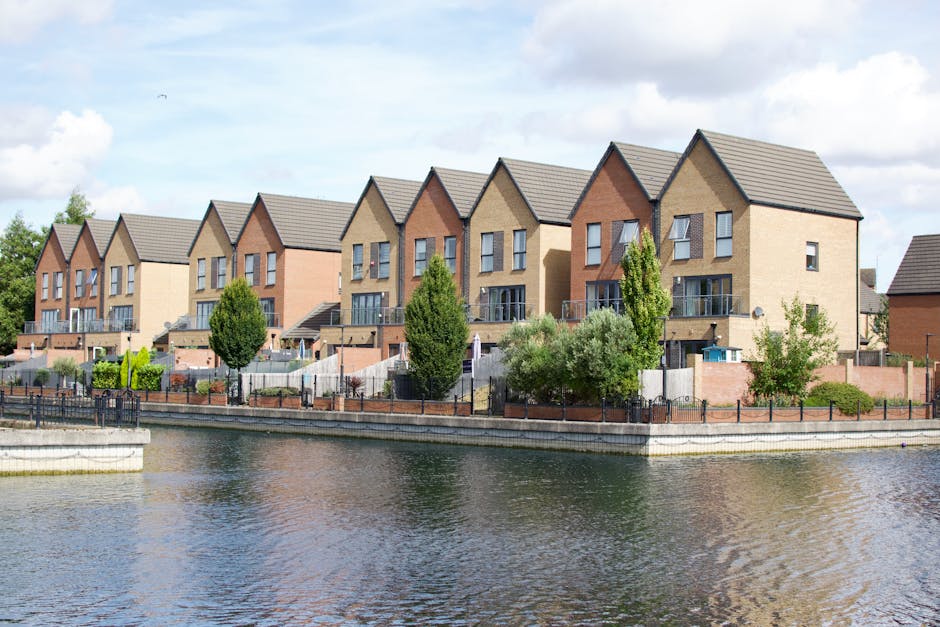 A row of modern multi-storey residential houses with brick and beige facades, pitched roofs, and large windows, situated along a waterfront on a sunny day with partly cloudy skies. The properties feature small front gardens enclosed by low metal fences, with a variety of trees and shrubs, including tall, narrow trees and bushy greenery. The houses are accessed via small outdoor stairs leading to doorways, and some have balconies with glass railings. A paved pathway runs parallel to the water, while the water itself reflects the buildings and the sky above. In the foreground, there is a concrete water edge with a metal railing. The scene appears to be part of a residential area that may require house removals or furniture transport services, such as those offered by Man and Van Enfield, especially for properties with tight access points and outdoor spaces for loading and unloading items.