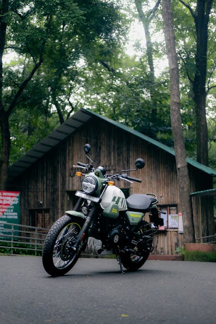 A black electric motorcycle with green and white accents is parked on a paved surface in front of a rustic wooden cabin surrounded by tall, leafy trees. The motorcycle features a round headlight, rearview mirrors, and a leather seat, with a small storage compartment mounted on the side. The scene is illuminated by natural daylight filtering through the forest canopy. The cabin's wooden exterior has a sloped roof and appears to be part of a rural property, with a notice board visible on the wall. This image relates to house removals and relocation services provided by Man and Van Enfield, illustrating the careful handling and transportation of valuable items such as motorcycles during home relocation or furniture transport in Enfield.