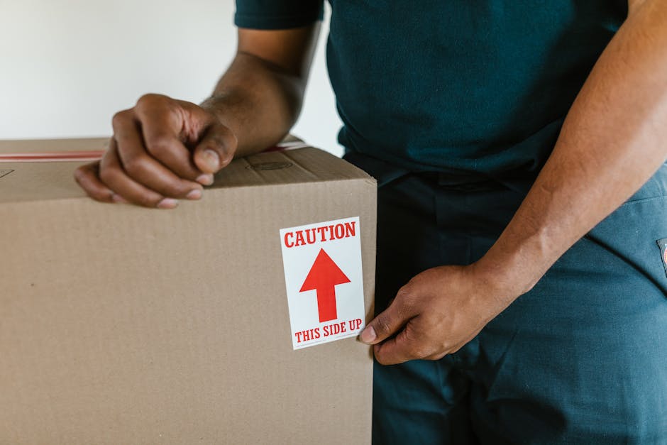 A person wearing a dark T-shirt and blue work pants is holding a large cardboard moving box with both hands, ensuring the top side remains up, as indicated by a red and white caution label that reads 'CAUTION THIS SIDE UP' with an upward arrow. The scene appears to take place inside a property, possibly near an entrance or doorway, with natural lighting illuminating the workspace. The cardboard box is unsealed and appears to be ready for transport as part of a home relocation or furniture transport process. The individual is preparing the box for safe handling during packing and moving activities. This image reflects the loading process, with a focus on proper handling and safety considerations during household removals, fitting within the context of furniture and belongings being organized for transportation by a professional removals service like Man and Van Enfield.